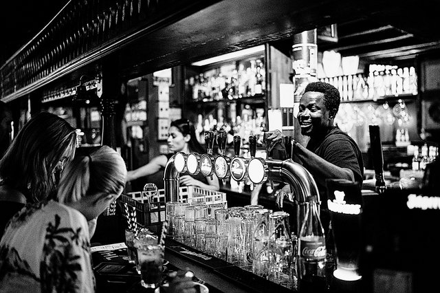 two women sit in front of the beer taps at the bar of a pub, while the bar tender pulls a pint
